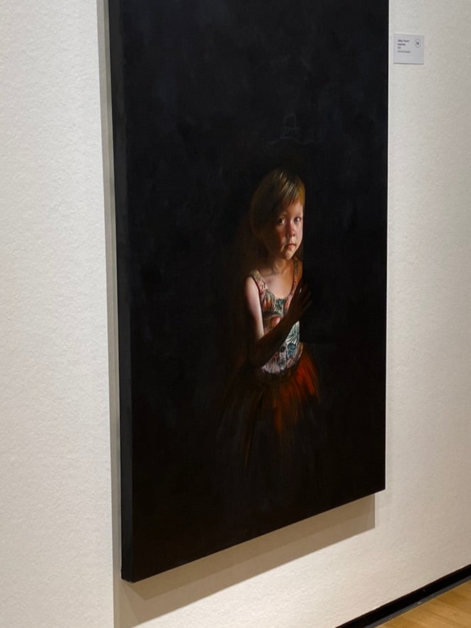 Das Bild mit dem Titel Augenhöhe von Tatjana Kpunkt im Museum Biberach, ein Porträt eines kleinen Mädchens, Lebensgroß auf schwarzem Hintergrund von einem einzelnen Lichtstrahl beleuchtet. Das Gemälde auf Leinwand ist 70x140 cm groß und das Mädchen derart positioniert, dass sie auf realer Augenhöhe auf dem Bild vorzufinden ist.